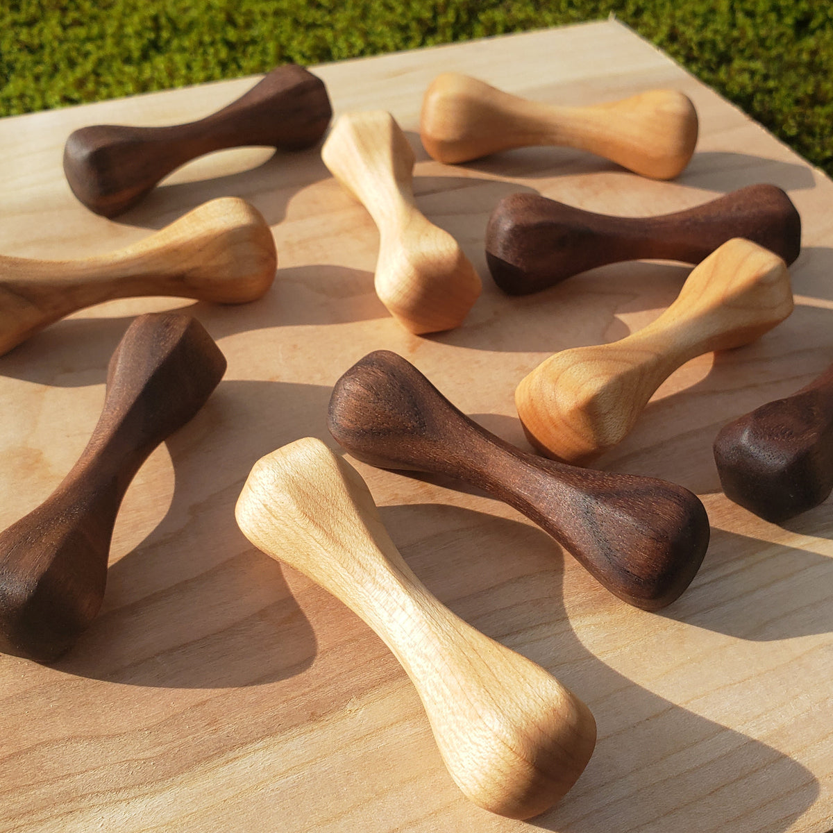 Ten handcrafted wooden knuckle roller pocket sculptures carved out of maple and walnut wood, arranged on a square light wood board outdoors on green moss.
